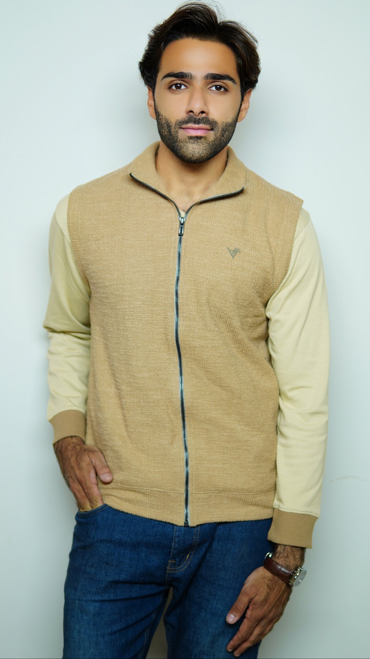 Man wearing a beige zip-up jacket and blue jeans against a light gray background